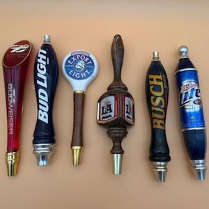 May include: A collection of six beer tap handles. The handles feature various designs and logos, including "Budweiser," "Bud Light," "Export Light," "Busch Beer," and "Miller Lite." The handles are made of various materials, including wood, metal, and plastic.