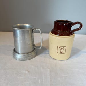 May include: Two vintage drinking vessels. A silver-colored metal beer mug with a handle and a base. Next to it is a cream-colored ceramic jug with a brown top and handle, featuring a brown bunny logo.