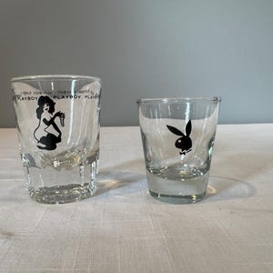 May include: Two clear shot glasses. One features a black silhouette of a woman and the word "PLAYBOY" in a repeating pattern. The other glass has a black bunny logo with a bow tie.