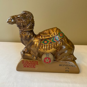 May include: A gold ceramic camel figurine with a colorful saddle and a red star on a gold base. The base reads "A.A.O.N.M. Shrine" and "U.S. Bicentennial 1776 1976".