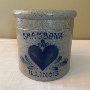 Puede incluir: Un tarro de cerámica gris con letras azules que dicen "Shabbona, Illinois". El tarro tiene un corazón azul con un diseño de remolino azul a cada lado del corazón.