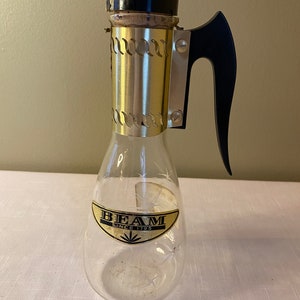 May include: A clear glass decanter with a gold metal top and a black plastic handle. The decanter has a label that reads "BEAM SINCE 1795".