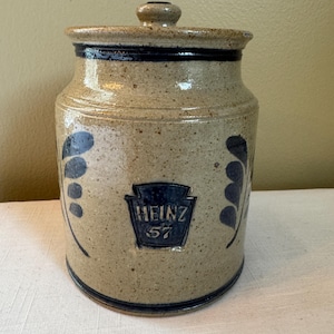 Peut inclure: Une cruche en grès avec un couvercle, décorée de peinture bleue et blanche. La cruche porte un logo Heinz 57 et un motif de feuilles.