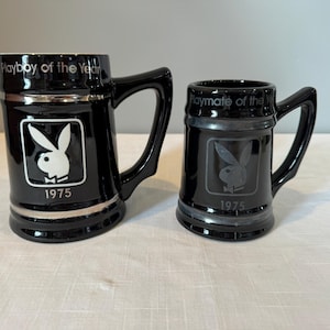 May include: Two black ceramic mugs with silver trim and handles. The larger mug reads "Playboy of the Year 1975" and features the Playboy bunny logo. The smaller mug reads "Playmate of the Year 1975" with the same logo.