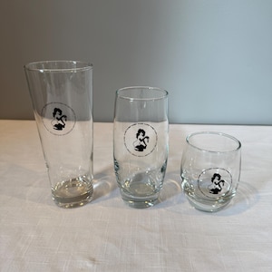 May include: Three clear drinking glasses of varying sizes. Each glass has a circular black and white logo. The glasses are on a white surface with a gray background. The glasses are empty.
