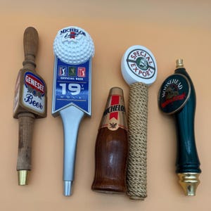 May include: A collection of beer tap handles with various designs. One handle features a golf ball, another has "Genesee Beer" text, and a third has "Moosehead" branding. Other handles include "Michelo" and "Special Export" branding.