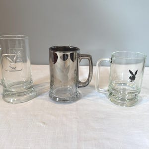 May include: Three clear glass beer mugs with handles. The mugs feature the Playboy bunny logo in different styles. One mug has the logo embossed, another has a black graphic, and the third has a silver metallic finish.
