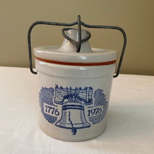Puede incluir: Tarro de cerámica blanco con un diseño de Campana de la Libertad azul y blanco. El tarro tiene un asa de alambre y un borde rojo alrededor de la tapa. El tarro está decorado con las fechas 1776 y 1976.