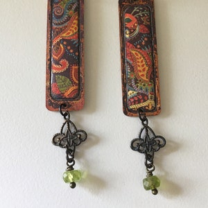 May include: A pair of dangle earrings with a rectangular copper frame and a colorful paisley pattern. The earrings have a green glass bead and a decorative bronze charm.