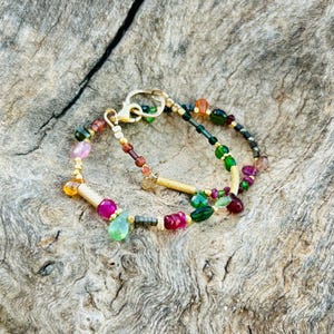 Puede incluir: Una colorida pulsera de cuentas con detalles dorados. La pulsera presenta una variedad de piedras preciosas en tonos verdes, rosas, rojos y naranjas. El cierre es dorado. La pulsera se muestra sobre una superficie de madera.