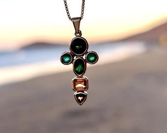 May include: A silver pendant necklace featuring a cross-shaped design. The pendant is adorned with a combination of green and orange gemstones. The necklace is suspended from a delicate silver chain, creating an elegant and eye-catching piece of jewelry.