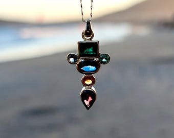 May include: A pendant necklace with a silver chain. The pendant features a cross-like design with various colorful gemstones, including green, blue, orange, and pink. The gemstones are set in a silver metal setting. The background is blurred, suggesting an outdoor setting.