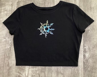 May include: Black crop top with a crew neck and short sleeves. The shirt has a colourful sunburst design on the front, with a blue centre and yellow and blue accents. The design has a tribal or celestial aesthetic.