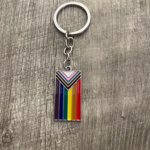 May include: A silver keychain with a rainbow-colored enamel charm. The charm features a chevron design with seven stripes in the colors of the rainbow flag.
