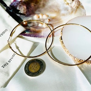 Puede incluir: Dos aros de oro sobre una superficie blanca con una concha de nácar y una pequeña moneda. Los aros son grandes y tienen un diseño simple. El texto "ZH2 STUDIO" es visible en una bolsa de tela blanca.