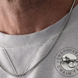 May include: A silver chain necklace with a small round tag that says "PB" on it. The tag is attached to the chain with a lobster clasp. The chain is on a person's neck and the tag is hanging down in front of the person's chest. The tag is surrounded by a circle that says "Engravable Chain Tag".