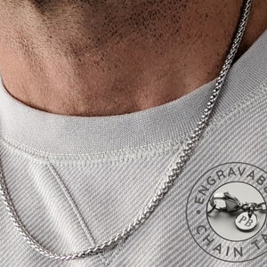 May include: A silver chain necklace with a small round tag that says "PB" on it. The tag is attached to the chain with a lobster clasp. The chain is on a person's neck and the tag is hanging in the centre of the chest. The text around the tag says "Engravable Chain Tag".