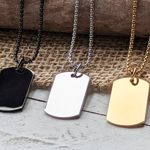 May include: Three silver dog tag necklaces with different finishes: black, silver, and gold. The tags are blank and have a rounded rectangular shape.