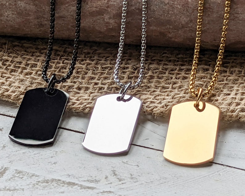 May include: Three silver dog tag necklaces with different finishes: black, silver, and gold. The tags are blank and have a rounded rectangular shape.