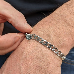 Puede incluir: Una pulsera de cadena plateada con un peque&ntilde;o dije redondo grabado con las letras "A&B". La pulsera est&aacute; hecha de eslabones ovalados entrelazados y se abrocha en una mu&ntilde;eca.