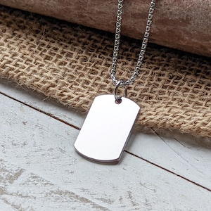 May include: A silver dog tag necklace with a blank rectangular tag. The tag has rounded corners and a small hole at the top. The necklace is on a light brown burlap surface.