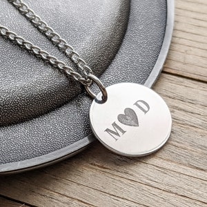 Peut inclure: Collier en argent avec un pendentif rond gravé des lettres "M" et "D" séparées par un symbole de cœur.