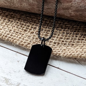 May include: A black dog tag necklace with a black chain. The dog tag is blank and has a rounded rectangular shape.