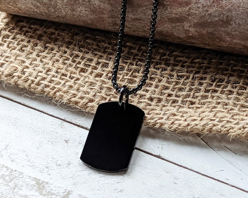 May include: A black dog tag necklace with a black chain. The dog tag is blank and has a rounded rectangular shape.