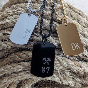 May include: Three silver, black, and gold dog tag necklaces with different engravings. The silver tag has the number 85 and a hockey stick and puck design. The black tag has two crossed pickaxes and the number 87. The gold tag has the initials "DR".