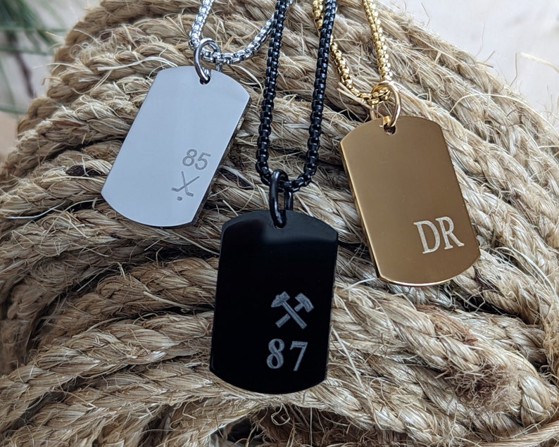 May include: Three silver, black, and gold dog tag necklaces with different engravings. The silver tag has the number 85 and a hockey stick and puck design. The black tag has two crossed pickaxes and the number 87. The gold tag has the initials "DR".