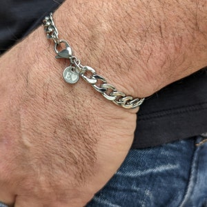 Puede incluir: Una pulsera de cadena de plata con cierre de mosquet&oacute;n y un peque&ntilde;o dije redondo. El dije tiene grabada la letra "E". La pulsera se lleva en la mu&ntilde;eca, con una camisa oscura y vaqueros azules de fondo.