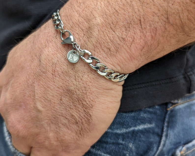 Puede incluir: Una pulsera de cadena de plata con cierre de mosquet&oacute;n y un peque&ntilde;o dije redondo. El dije tiene grabada la letra "E". La pulsera se lleva en la mu&ntilde;eca, con una camisa oscura y vaqueros azules de fondo.