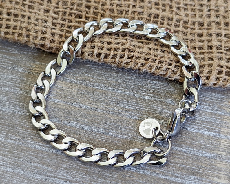 Puede incluir: Una pulsera de cadena de plata con cierre de mosquet&oacute;n. La pulsera tiene un acabado pulido y un peque&ntilde;o dije redondo con un coraz&oacute;n y la letra "E". La pulsera se exhibe sobre una superficie de madera.