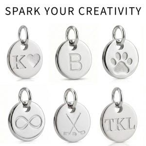 Puede incluir: Colgantes redondos plateados con varios dise&ntilde;os grabados. Uno muestra la letra "K" con un coraz&oacute;n, otro la letra "B", y un tercero una huella de pata. La imagen tambi&eacute;n incluye colgantes con un s&iacute;mbolo de infinito, palos de hockey y las iniciales "TKL". El texto "SPARK YOUR CREATIVITY" est&aacute; en la parte superior.