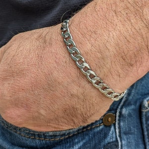 Puede incluir: Una pulsera de cadena de plata se lleva en la mu&ntilde;eca. La pulsera est&aacute; hecha de eslabones entrelazados. La persona lleva vaqueros azules y una camisa oscura. La pulsera es una pieza de joyer&iacute;a cl&aacute;sica.