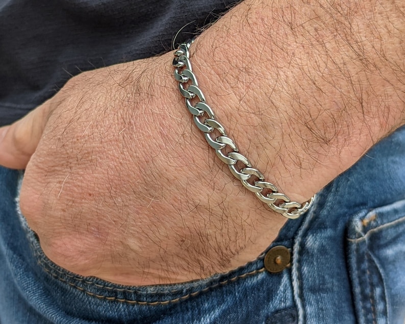 Puede incluir: Una pulsera de cadena de plata se lleva en la mu&ntilde;eca. La pulsera est&aacute; hecha de eslabones entrelazados. La persona lleva vaqueros azules y una camisa oscura. La pulsera es una pieza de joyer&iacute;a cl&aacute;sica.
