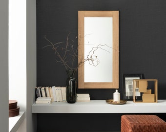 May include: A white shelf with a wooden framed mirror, a vase with dried branches, a stack of books, a small gold metal sculpture, and a brown leather ottoman. The shelf is mounted on a black wall.