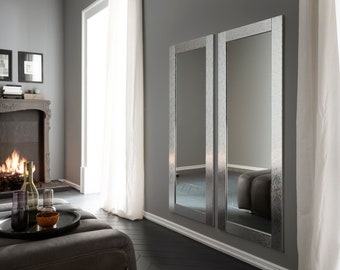 May include: Two large, silver-framed mirrors hang on a gray wall in a modern living room. The mirrors reflect the room's interior, including a fireplace and a couch.