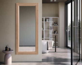 May include: A rectangular, light-colored wooden framed mirror is mounted on a neutral-toned wall. The mirror is the focal point of a modern bathroom, with a neutral color palette. A small side table and a rug are in the foreground.