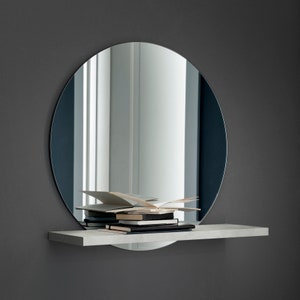 May include: A round, frameless mirror with a gray shelf below. The shelf holds several books and papers. The mirror reflects a bright, white interior space. The background is a dark gray wall.