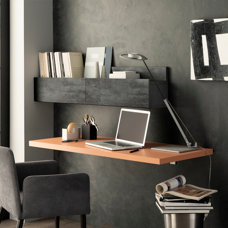 Pu&ograve; includere: Una scrivania moderna per ufficio domestico con uno scaffale a parete e un computer portatile. La scrivania &egrave; in legno e ha una finitura marrone chiaro. Lo scaffale &egrave; in legno grigio scuro e ha due scomparti. Il computer portatile &egrave; aperto e ha uno schermo vuoto. C'&egrave; una lampada sulla scrivania e una pila di libri sul pavimento.