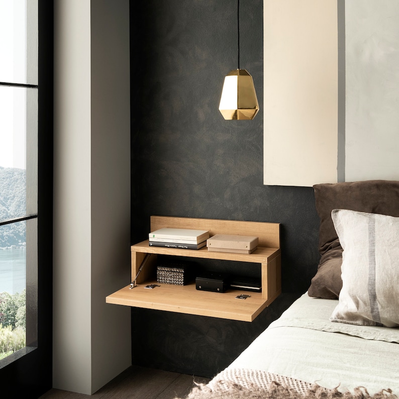 Pu&ograve; includere: Un ripiano a muro in legno con un fronte a ribalta. Il ripiano ha due scomparti e contiene alcuni libri e piccole scatole. Il ripiano &egrave; montato su una parete grigio scuro accanto a un letto con lenzuola bianche e una lampada a sospensione dorata.