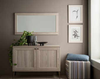 May include: A light wood cabinet with two doors and gold legs. The cabinet is decorated with a mirror, a potted plant, and decorative objects. The cabinet is in front of a light brown wall with two framed black and white prints.