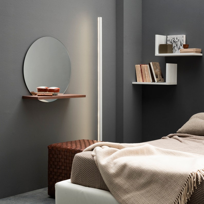 K&ouml;nnte beinhalten: Ein Schlafzimmer mit einem wei&szlig;en Bett, einem braunen geflochtenen Hocker und einem Wandspiegel mit einem Regal. Die Wand ist grau gestrichen und es gibt wei&szlig;e Regale an der Wand mit B&uuml;chern und Dekoartikeln.
