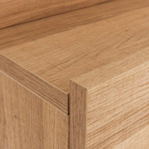 Pu&ograve; includere: Primo piano dell'angolo di un armadio in legno con finitura marrone chiaro. L'armadio ha un design semplice con una superficie liscia.