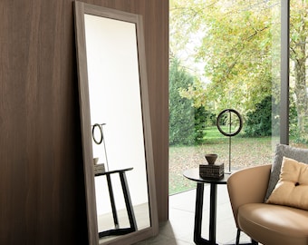 May include: A full-length mirror with a wood frame, reflecting a side table with a vase and a small object. The mirror is leaning against a wood wall, and a chair with a throw pillow is in the foreground.