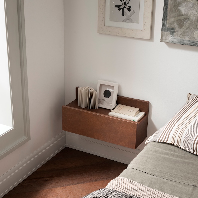 Pu&ograve; includere: Un comodino sospeso marrone con un libro, una piccola foto incorniciata e un libro con una copertina bianca in cima. Il comodino &egrave; montato su una parete bianca accanto a un letto con un cuscino a righe verde e bianco.