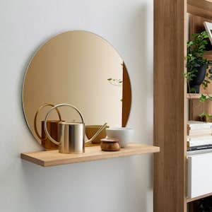 Op de afbeelding: Een houten plank met een ronde gouden spiegel, een gieter, een klein houten doosje en een witte kom. De plank is gemonteerd aan een witte muur.