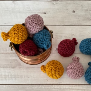 May include: A collection of eight crocheted balls in various colors, including pink, blue, yellow, and red, are displayed in a copper bucket and on a white wooden surface.