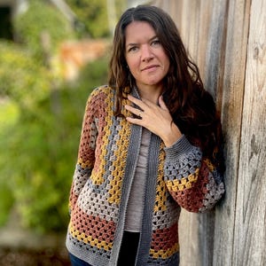 May include: A handmade crochet cardigan in a granny square pattern. The open-weave design features a color block of gray, mustard yellow, rust, and pink. The cardigan has long sleeves and a gray border.
